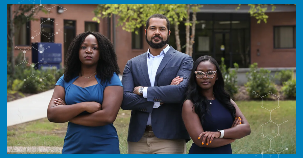 Two female and one male Black students standing outside on grass lawn in front of brick buildings.