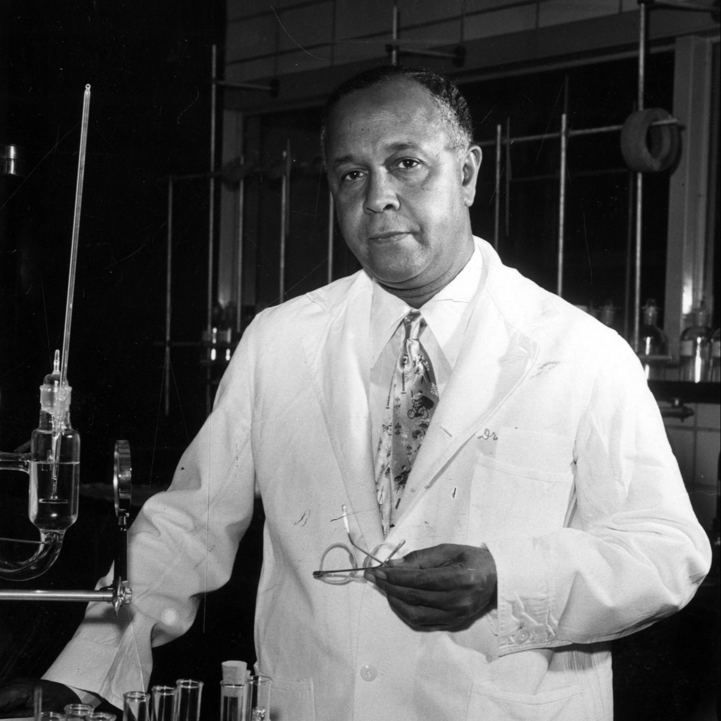 Black and white image of male in laboratory wearing white lab coat and holding glasses